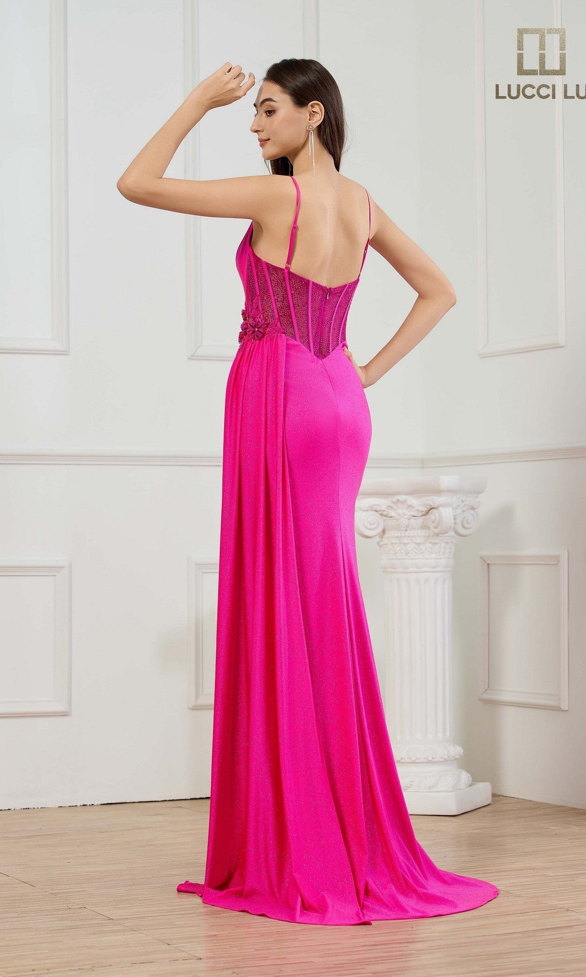 A woman stands with her back to the camera in an elegant room, wearing the Lucci Lu 1473—a bright pink, floor-length prom dress featuring thin straps, lace detailing, and delicate 3D floral appliques.